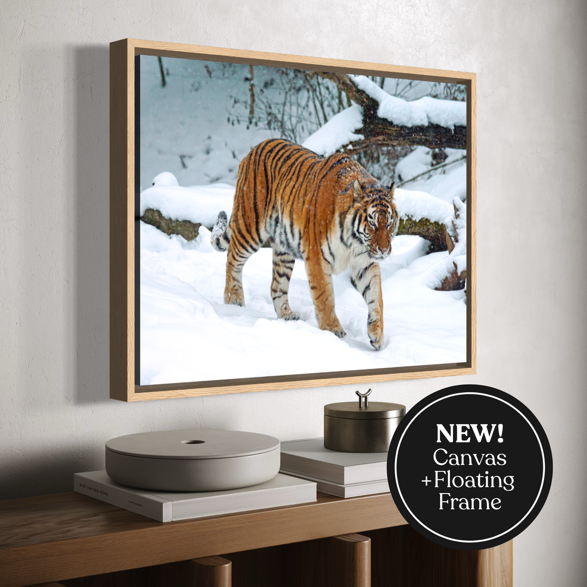 A framed canvas wall art print showing a tiger walking through a snowy forest, displayed above a wooden console table. The print has a light wood floating frame and a black circular badge in the lower right corner that reads 'NEW! Canvas + Floating Frame'.