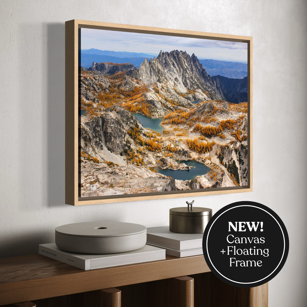 A framed canvas mountain landscape print hangs on a light gray wall above a wooden console table. The artwork shows rocky peaks, blue lakes, and autumn-colored trees.