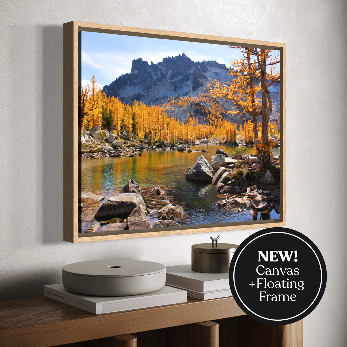 A framed canvas landscape wall art print showing a mountain, lake, and golden autumn trees, displayed on a wall above a console table. The print has a light wood floating frame, and a black circular badge in the lower right says 'NEW! Canvas + Floating Frame'.