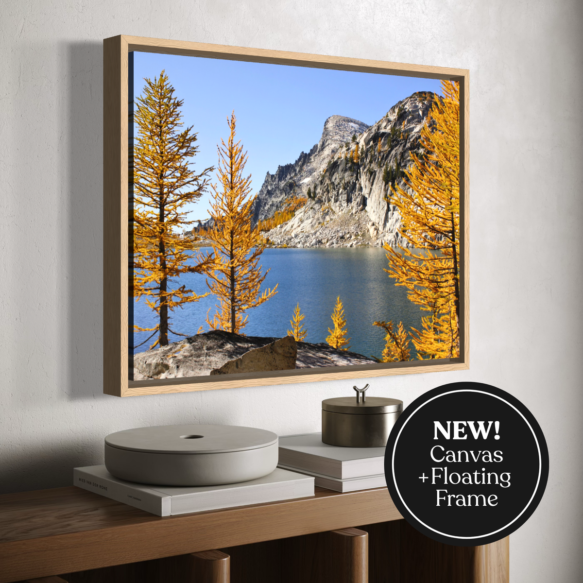A wooden framed landscape wall art print showing a mountain, lake, and golden trees, displayed on a wall above a wooden console with decorative objects. A black circular badge in the lower right says 'NEW! Canvas + Floating Frame'.