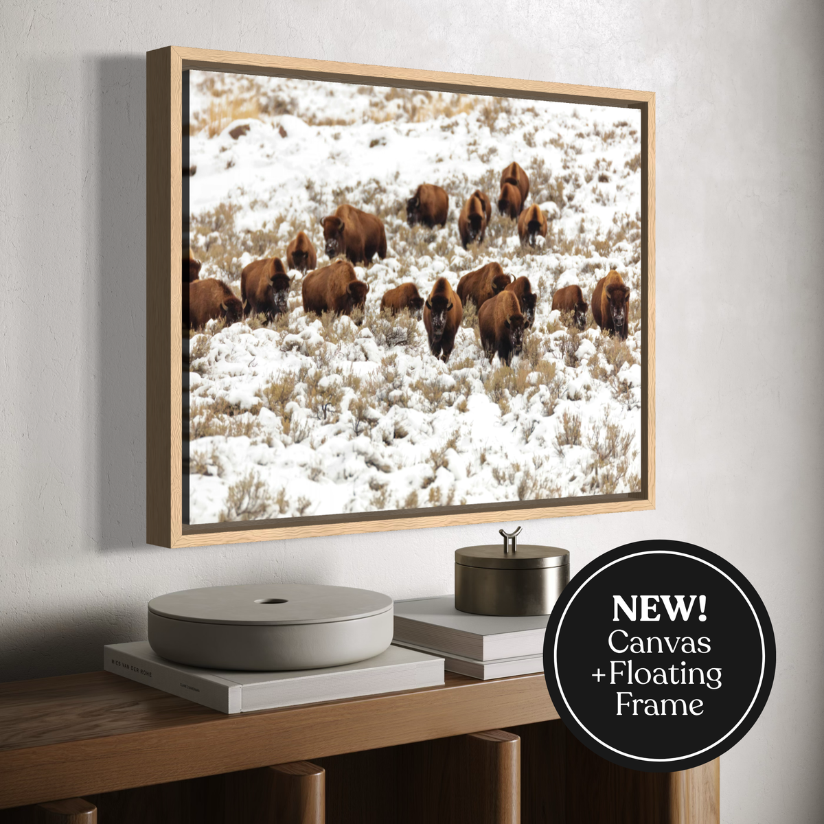 A framed canvas print of bison in a snowy field, displayed on a wall above a wooden console table with decorative objects. A black circular badge in the lower right corner reads 'NEW! Canvas + Floating Frame'.