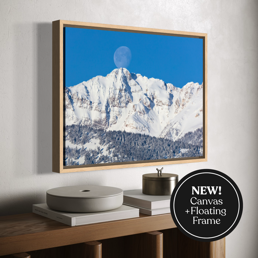 Framed canvas wall art showing a snowy mountain landscape under a bright blue sky with a moon visible, displayed in a floating frame above a console table. A black circular badge reads “NEW! Canvas + Floating Frame.