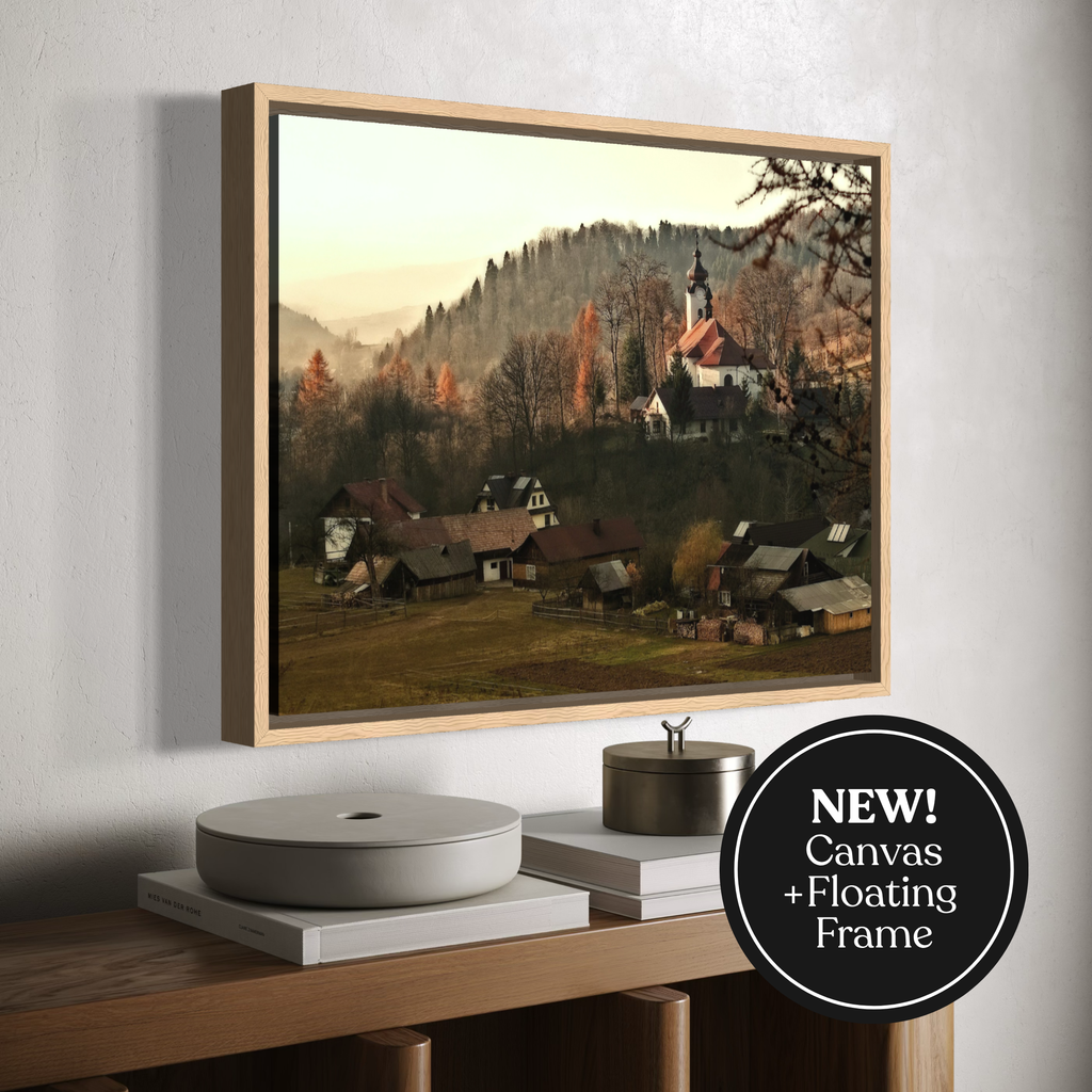 Framed wall art showing a mountain village scene with houses, trees, and a church, displayed above a wooden console. A black circular badge reads “NEW! Canvas + Floating Frame.