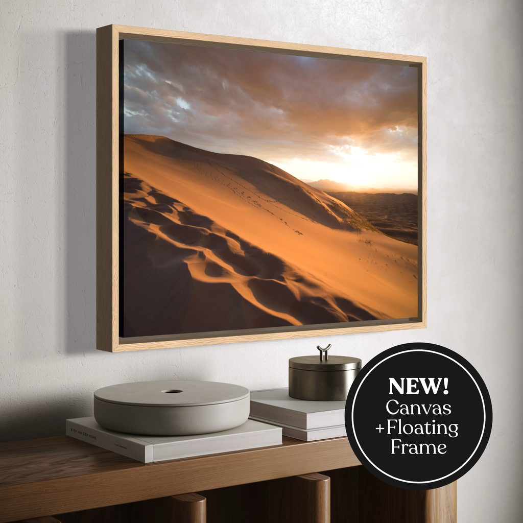 A framed canvas desert landscape wall art print hangs above a wooden console table with books and a small container. The artwork shows warm orange sand dunes under a dramatic sky, and a black circular badge in the lower right says 'NEW! Canvas + Floating Frame'.