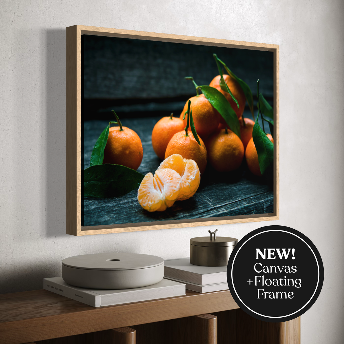 A framed wall art print showing a still life of oranges and green leaves on a dark wooden surface, displayed above a console table in a styled interior. A black circular badge in the lower right reads 'NEW! Canvas + Floating Frame'.