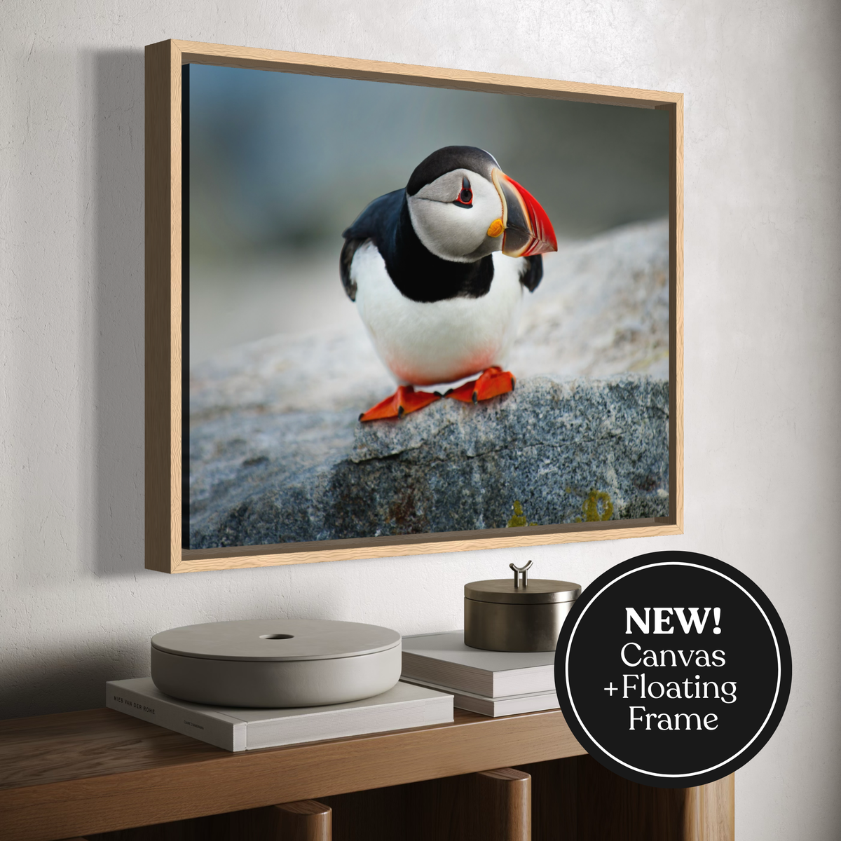 A framed canvas wall art print showing a puffin standing on a rock, displayed above a console table with books and decor. A black circular badge in the lower right says 'NEW! Canvas + Floating Frame'.