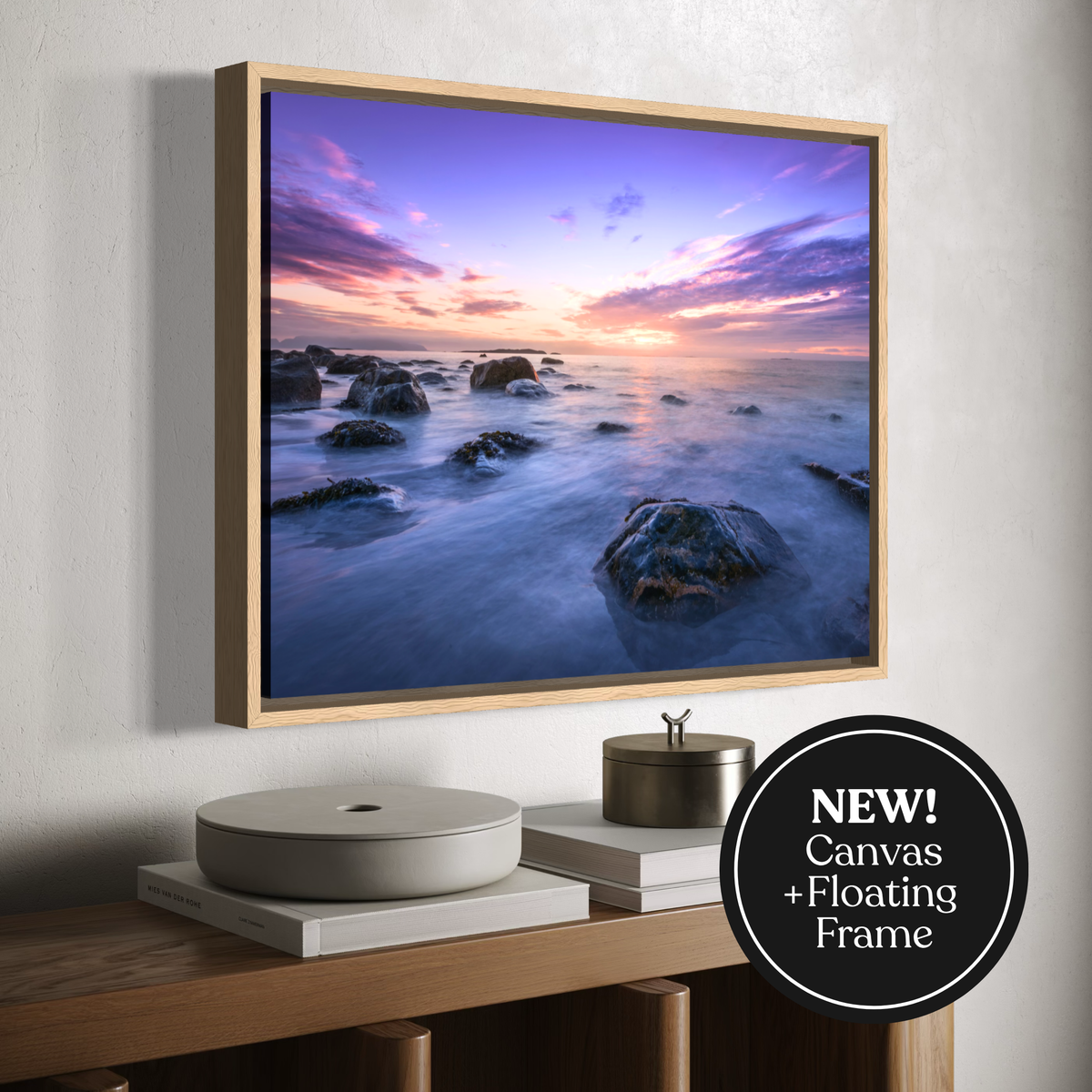A framed canvas wall art print showing a coastal seascape at sunset, mounted on a light wall above a wooden console with books and decor. A black circular badge in the lower right says 'NEW! Canvas + Floating Frame'.