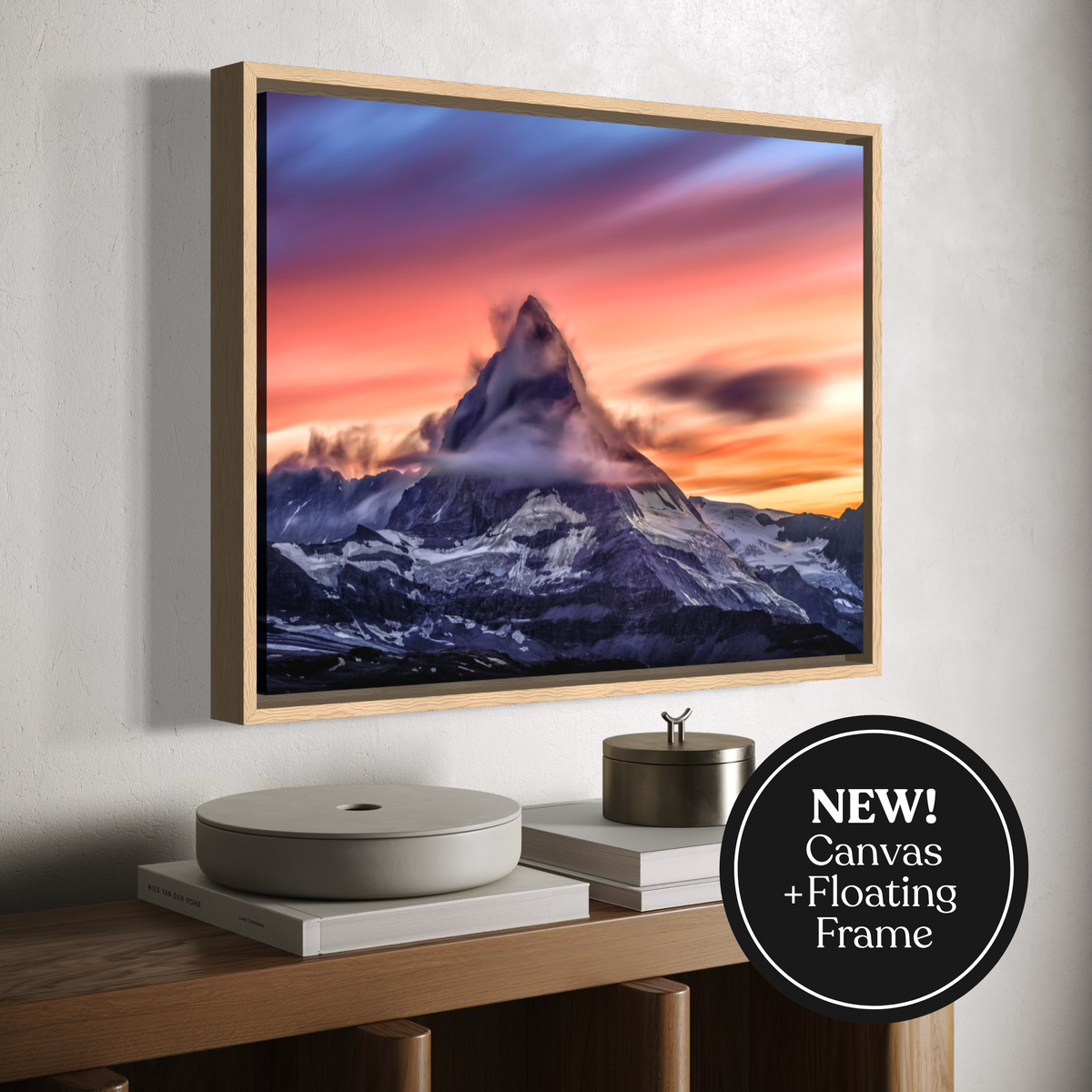 A framed canvas print of a mountain peak at sunset hangs on a light wall above a wooden console table with decorative objects. The artwork shows a snow-covered mountain under a vivid sky with purple, blue, pink, and orange tones.