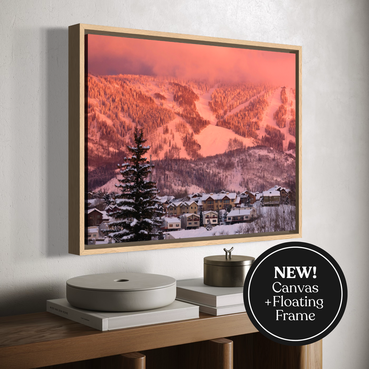 A framed canvas wall art print showing a snowy mountain village under a pink-orange sky, displayed above a wooden console with decorative objects. A black circular badge in the lower right says 'NEW! Canvas + Floating Frame'.
