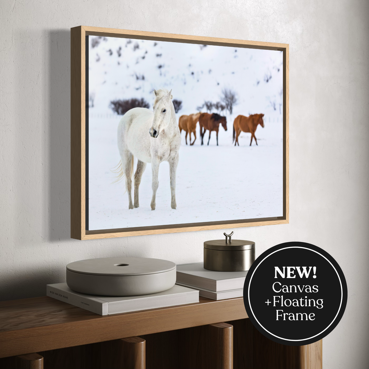 Framed canvas wall art showing a white horse standing in a snowy landscape with three brown horses in the background, displayed above a wooden console table. A black circular badge in the lower right reads 'NEW! Canvas + Floating Frame'.