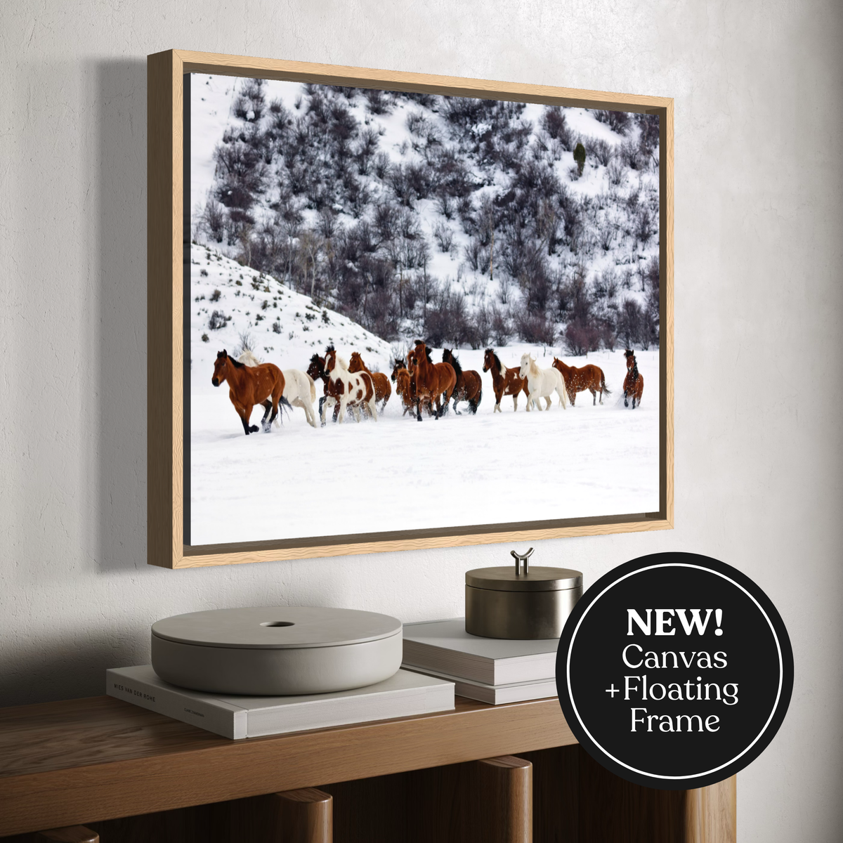 A framed canvas wall art print showing a herd of horses running through a snowy landscape, displayed above a wooden console table. The print has a light wood floating frame and a black circular badge in the lower right corner that reads 'NEW! Canvas + Floating Frame'.