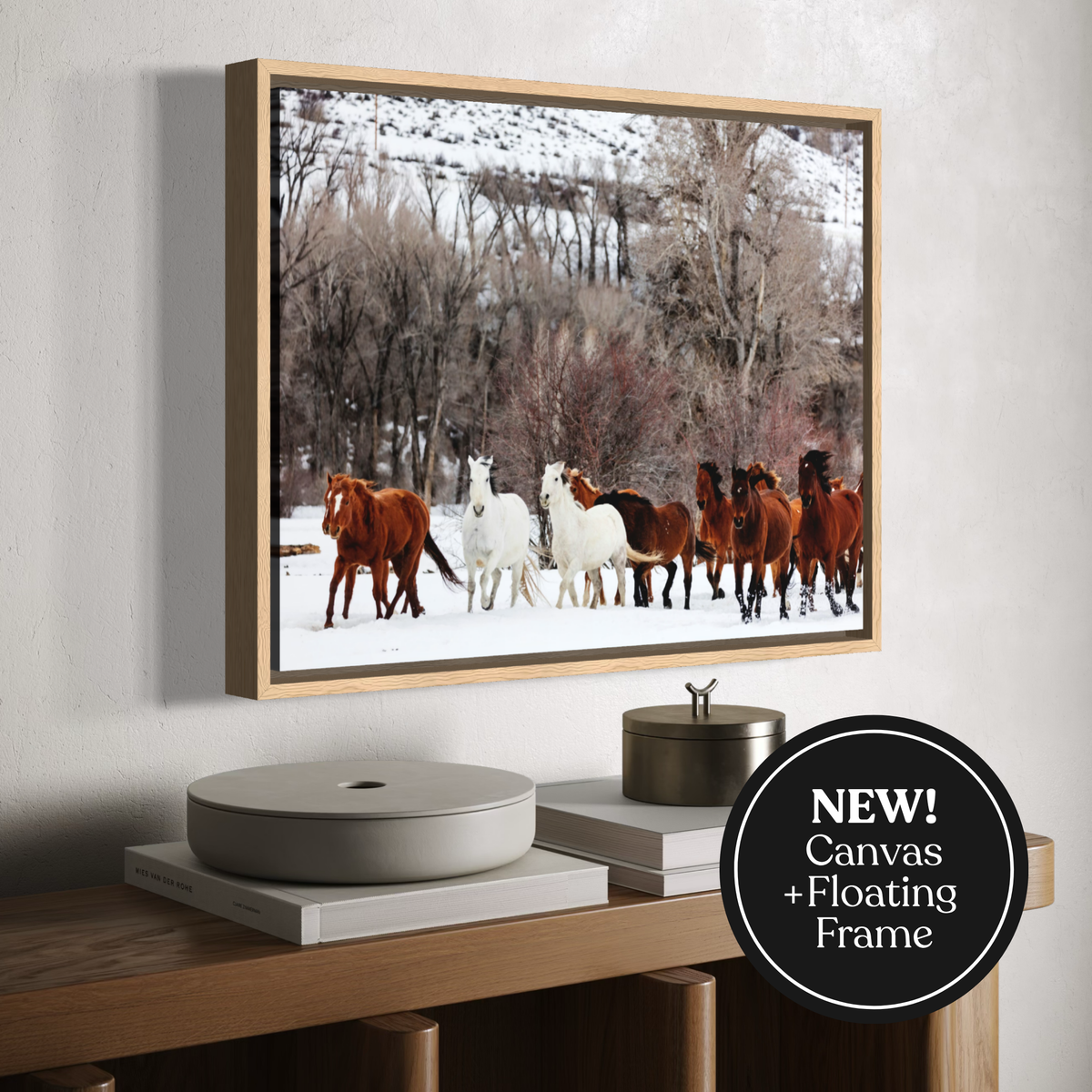 A framed canvas print of a herd of horses in a snowy landscape, styled on a wooden console table against a light gray wall. A black circular badge in the bottom right corner reads 'NEW! Canvas + Floating Frame'.