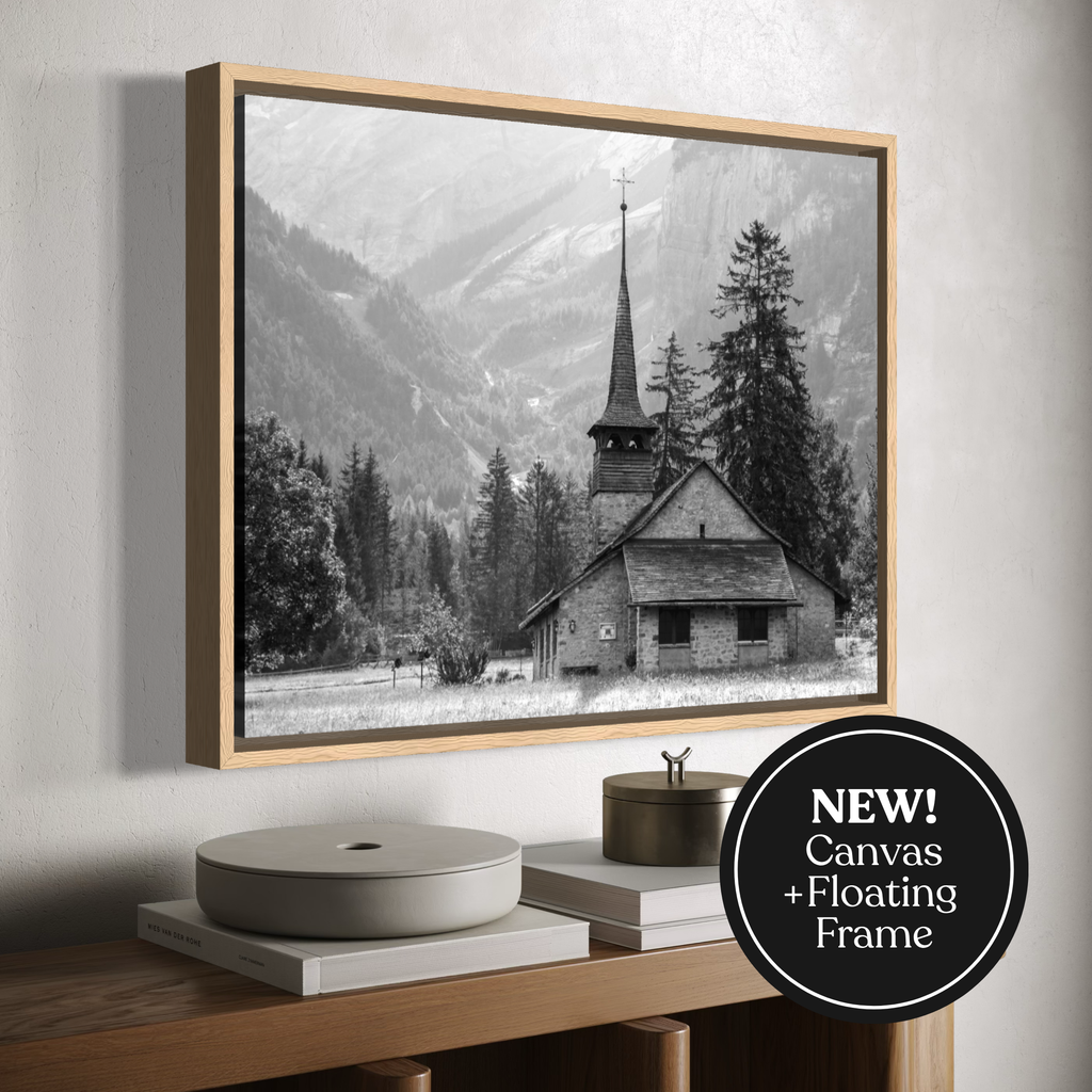 A framed canvas black-and-white landscape print of a mountain village with a church and trees, shown hanging on a light wall above a wooden console with books and decor. A circular badge in the lower right says 'NEW! Canvas + Floating Frame'.