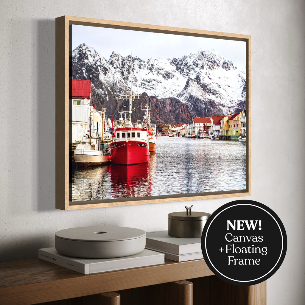A framed canvas wall art print showing a colorful harbor scene with boats, snowy mountains, and water, displayed on a wall above a wooden console. A black circular badge in the lower right says 'NEW! Canvas + Floating Frame'.