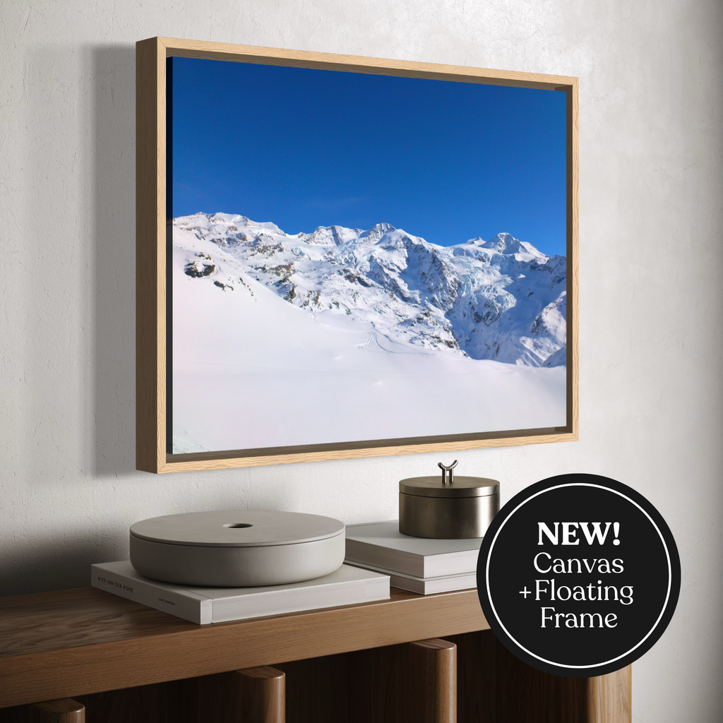 A framed mountain landscape print hangs on a light wall above a wooden console table. The artwork shows snow-covered peaks under a vivid blue sky, and the frame appears floating with a light wood border.