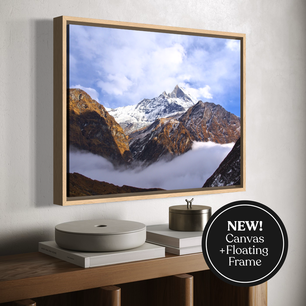 A framed canvas wall art print showing a mountain landscape with snow, clouds, and rocky peaks, displayed above a wooden console. The print is housed in a light wood floating frame.
