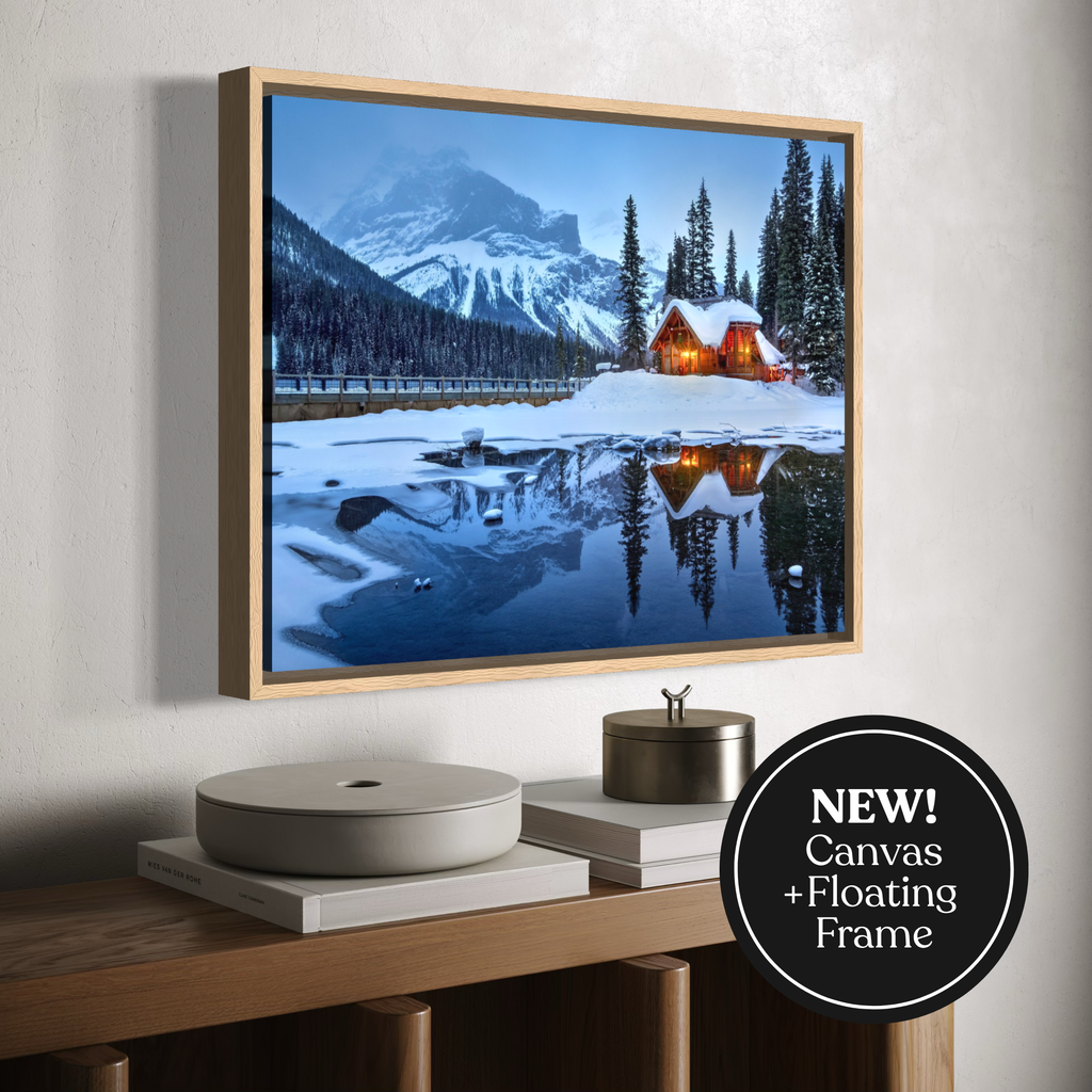 A framed canvas print of a snowy mountain landscape with a glowing cabin reflected in a lake, displayed on a wall above a wooden console. A black circular badge in the lower right reads 'NEW! Canvas + Floating Frame'.