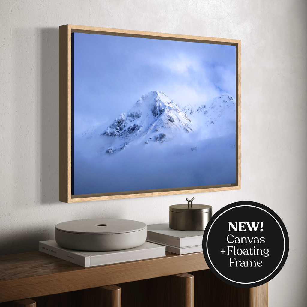 A framed canvas wall art print showing a snowy mountain peak rising through blue clouds, displayed above a wooden console with decorative objects. A black circular badge in the lower right reads “NEW! Canvas + Floating Frame.
