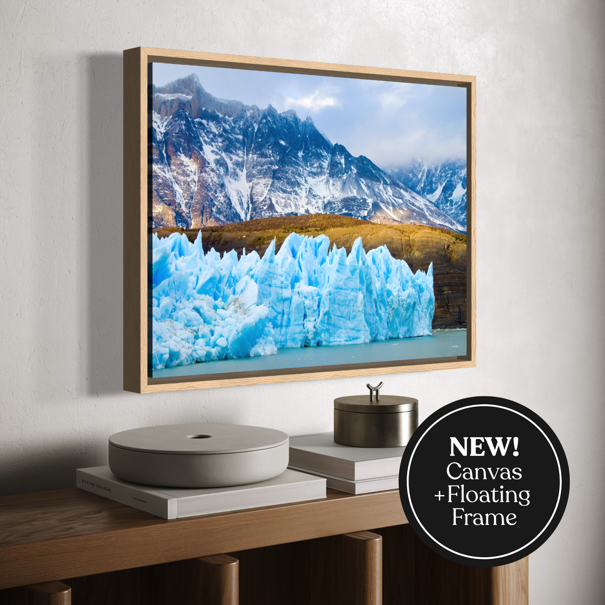 A framed wall art print showing a snowy mountain landscape with bright blue ice formations in the foreground, displayed above a wooden console. A black circular badge in the lower right reads 'NEW! Canvas + Floating Frame'.