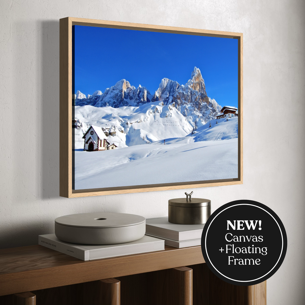 A framed canvas print of a snowy mountain landscape with a bright blue sky, displayed on a light gray wall above a wooden console table. The frame is shown floating at an angle, and a black circular badge in the bottom right corner reads 'NEW! Canvas + Floating Frame'.