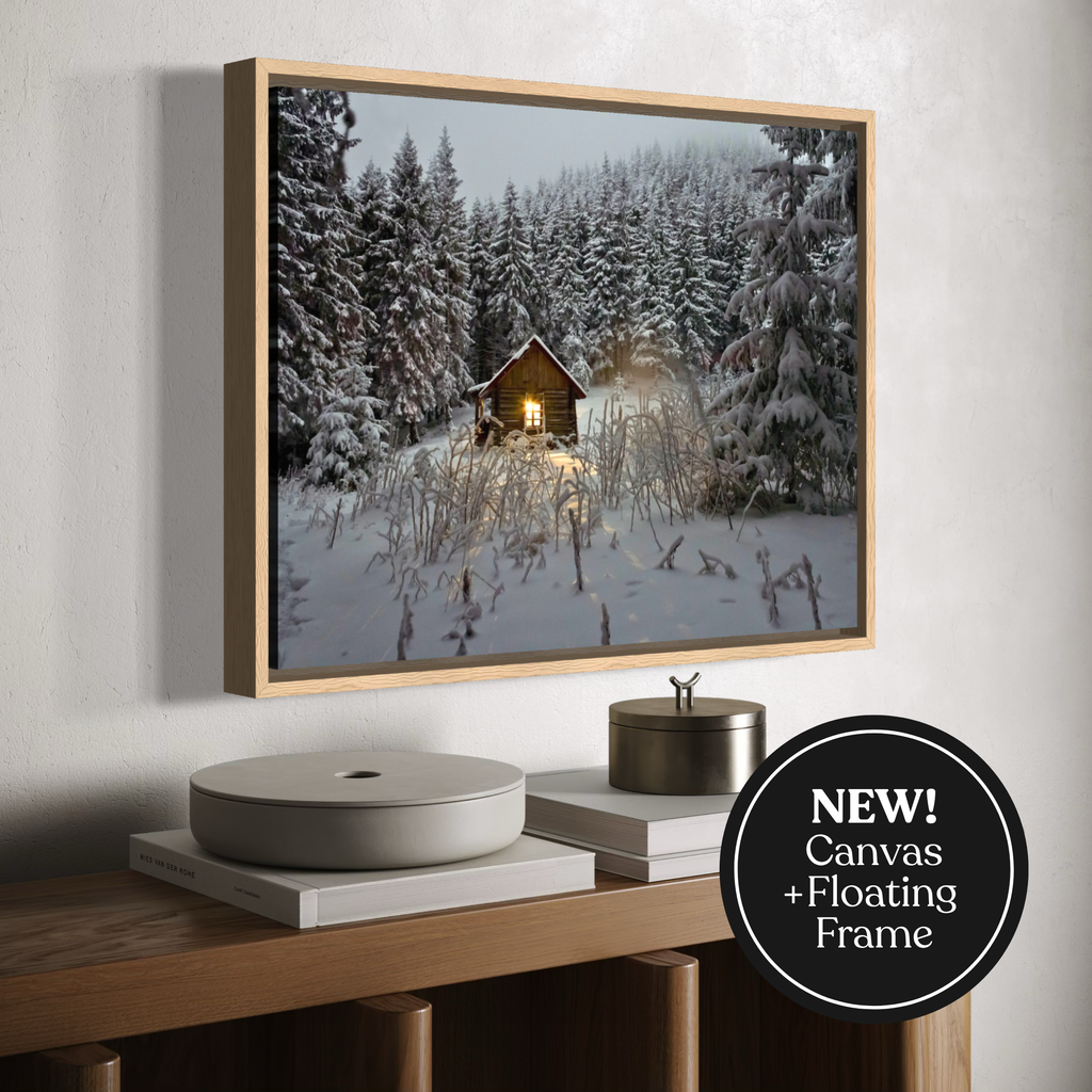 Framed canvas wall art showing a snowy forest cabin scene, displayed above a wooden console with books and decor. A black circular badge reads “NEW! Canvas + Floating Frame.