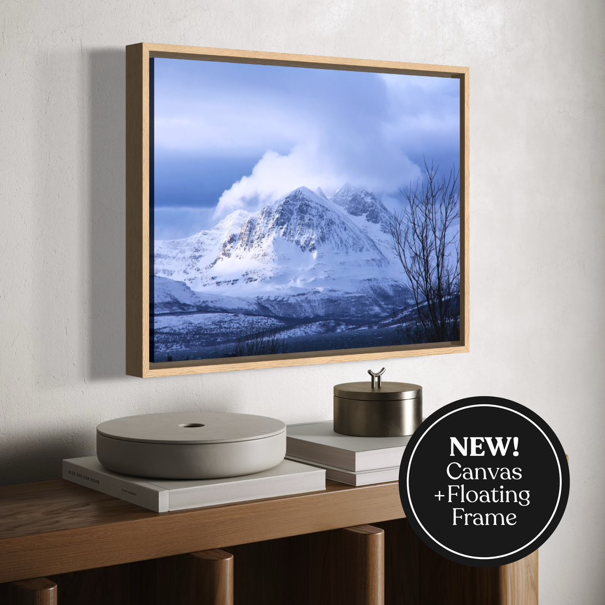 A framed wall art print showing a snowy mountain landscape under a blue cloudy sky, displayed above a wooden console with books and decor. A black circular badge in the lower right reads 'NEW! Canvas + Floating Frame'.