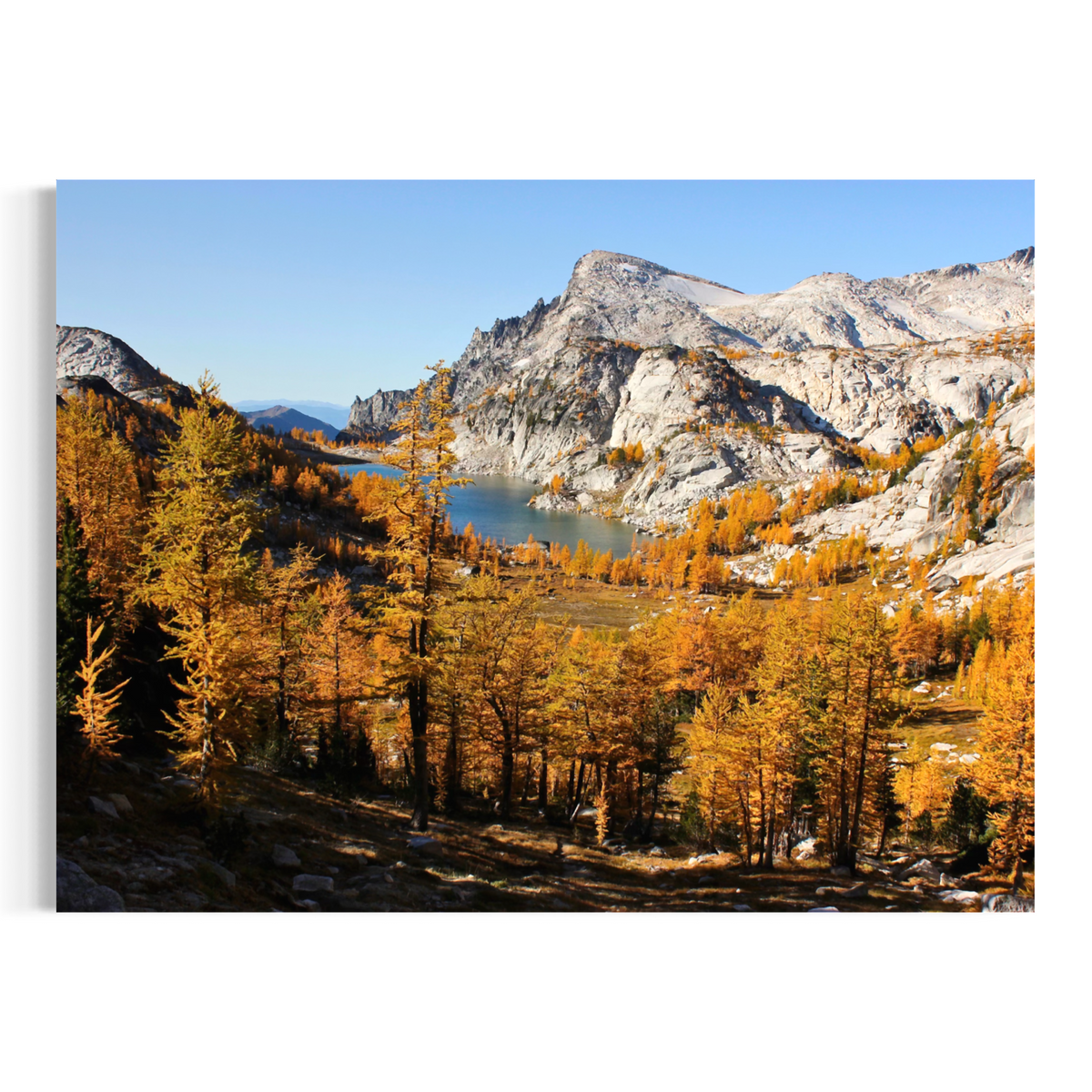 A wall art print or canvas showing a mountain landscape with a lake, rocky peaks, and golden autumn trees, displayed against a plain white background.