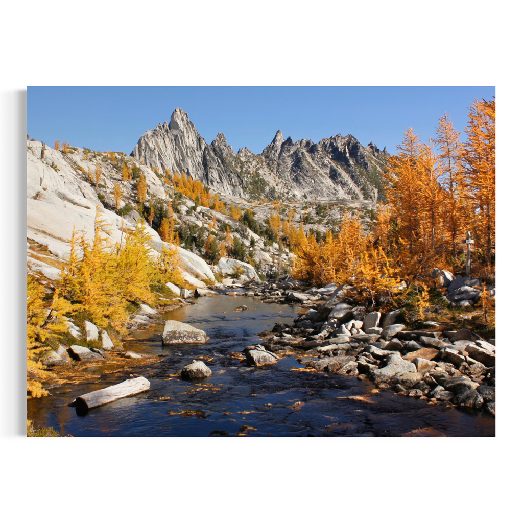 A rectangular wall art print or canvas showing a mountain landscape with jagged gray peaks, a rocky stream, and bright orange-yellow trees under a clear blue sky, displayed against a plain white background.