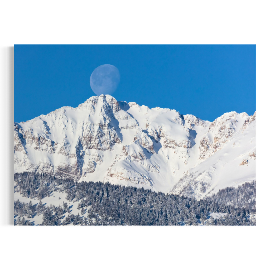 A wall art print or canvas showing a snowy mountain peak under a clear blue sky with a visible moon, photographed against a white wall.