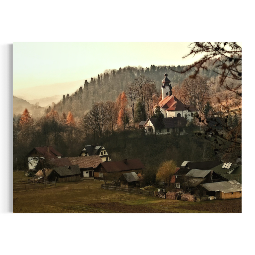 A scenic landscape photograph wall art print of a small mountain village with a church, farmhouses, and forested hills in the background, shown with a slight perspective tilt on a white background.