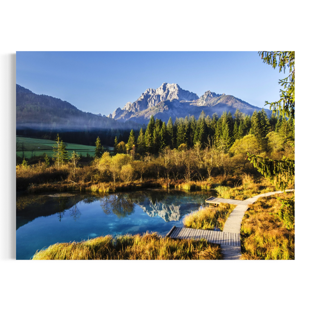 A rectangular wall art print or canvas showing a mountain landscape with a blue lake, evergreen forest, grassy marsh, and a wooden boardwalk in the foreground, photographed against a plain white background.
