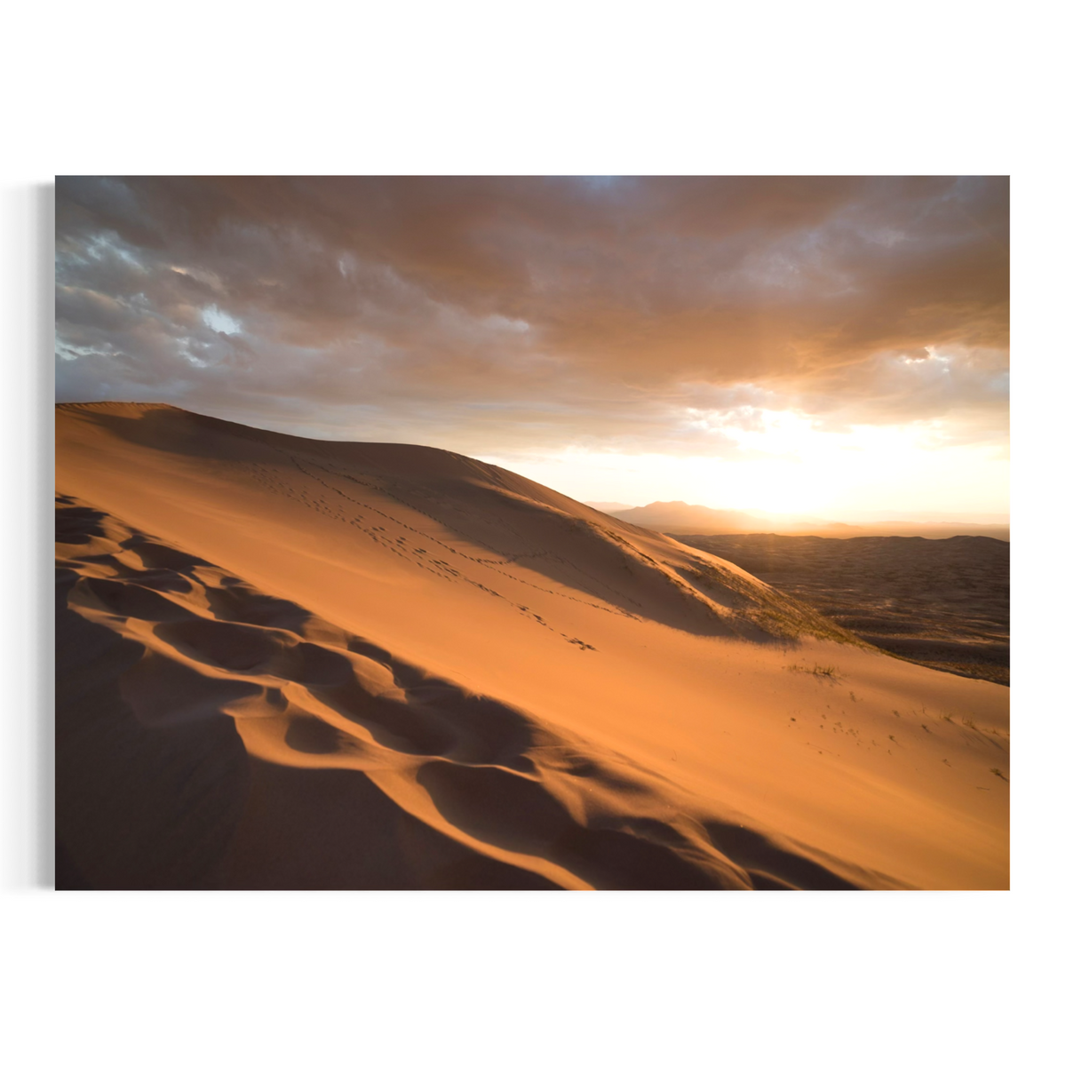 A rectangular wall art print showing a desert landscape at sunset, with golden sand dunes in the foreground, distant mountains, and dramatic clouds glowing orange and brown. The print is displayed against a clean white background.