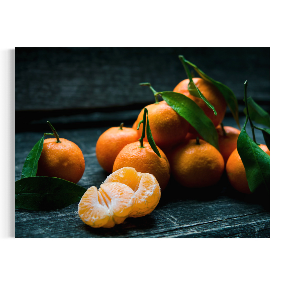 A photographic print of still life of several bright orange mandarins with green leaves, including one peeled mandarin showing the segmented white and orange flesh, arranged on a dark rustic wooden surface.