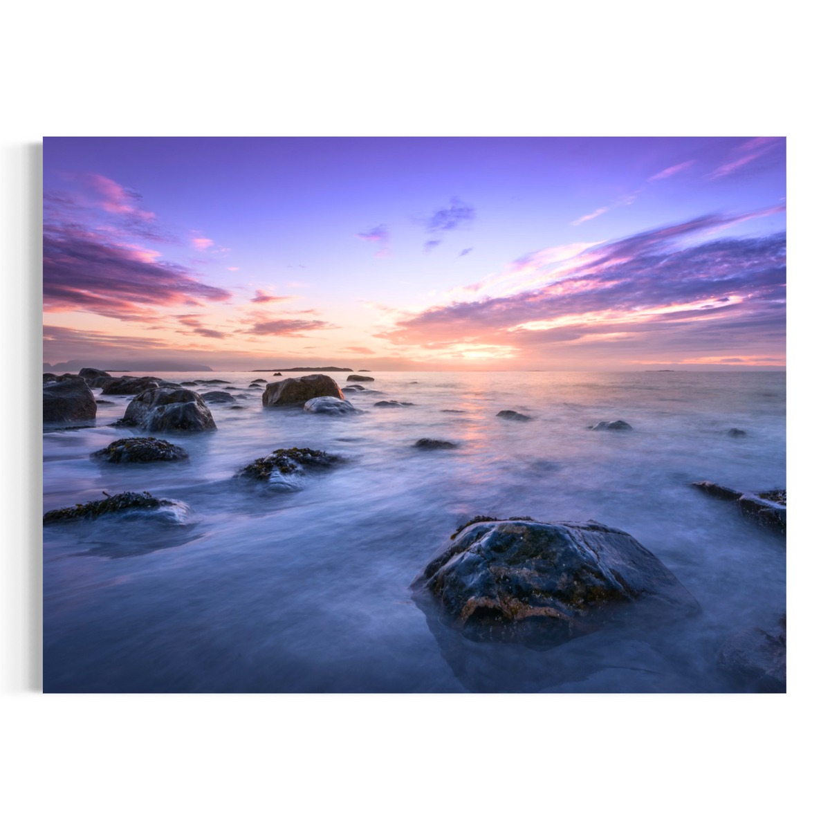 A rectangular wall art print showing a coastal seascape at sunset with purple and pink clouds, a glowing sun near the horizon, and dark rocks in the foreground, displayed against a light gray background.