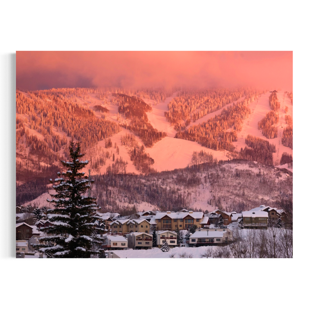 A rectangular wall art print showing a snowy mountain ski resort at sunset, with ski slopes, pine trees, and small buildings under a pink-orange sky, displayed against a white background.
