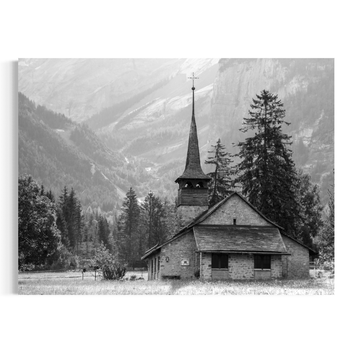 Black and white wall art print depicting a small mountain church with a tall steeple, surrounded by evergreen trees and rugged mountains in the background.