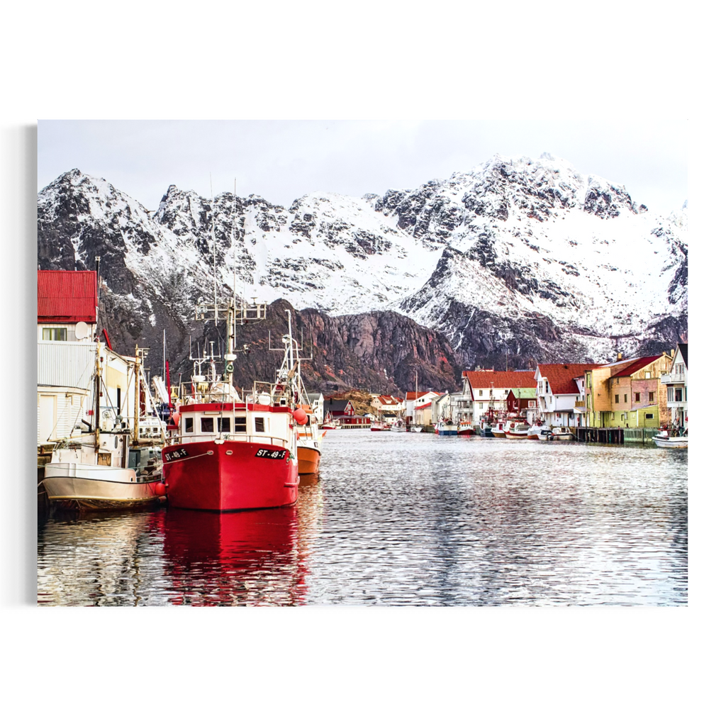 A wall art print or canvas showing a snowy mountain harbor scene with fishing boats in the foreground, colorful buildings along the water, and reflections in the water, presented against a plain white background.