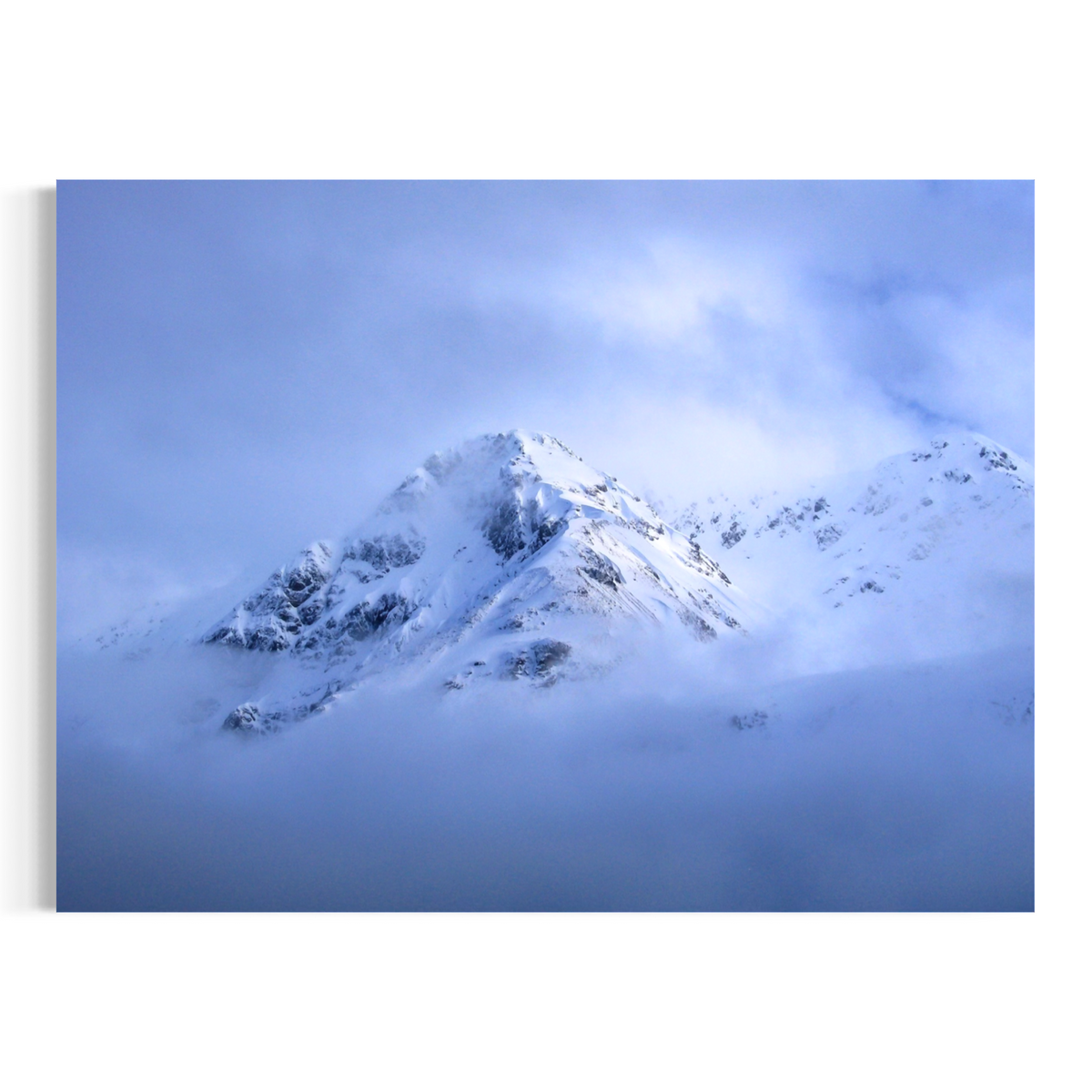 A wall art print or canvas showing a snow-covered mountain peak rising above low blue-gray clouds, displayed against a plain white wall.