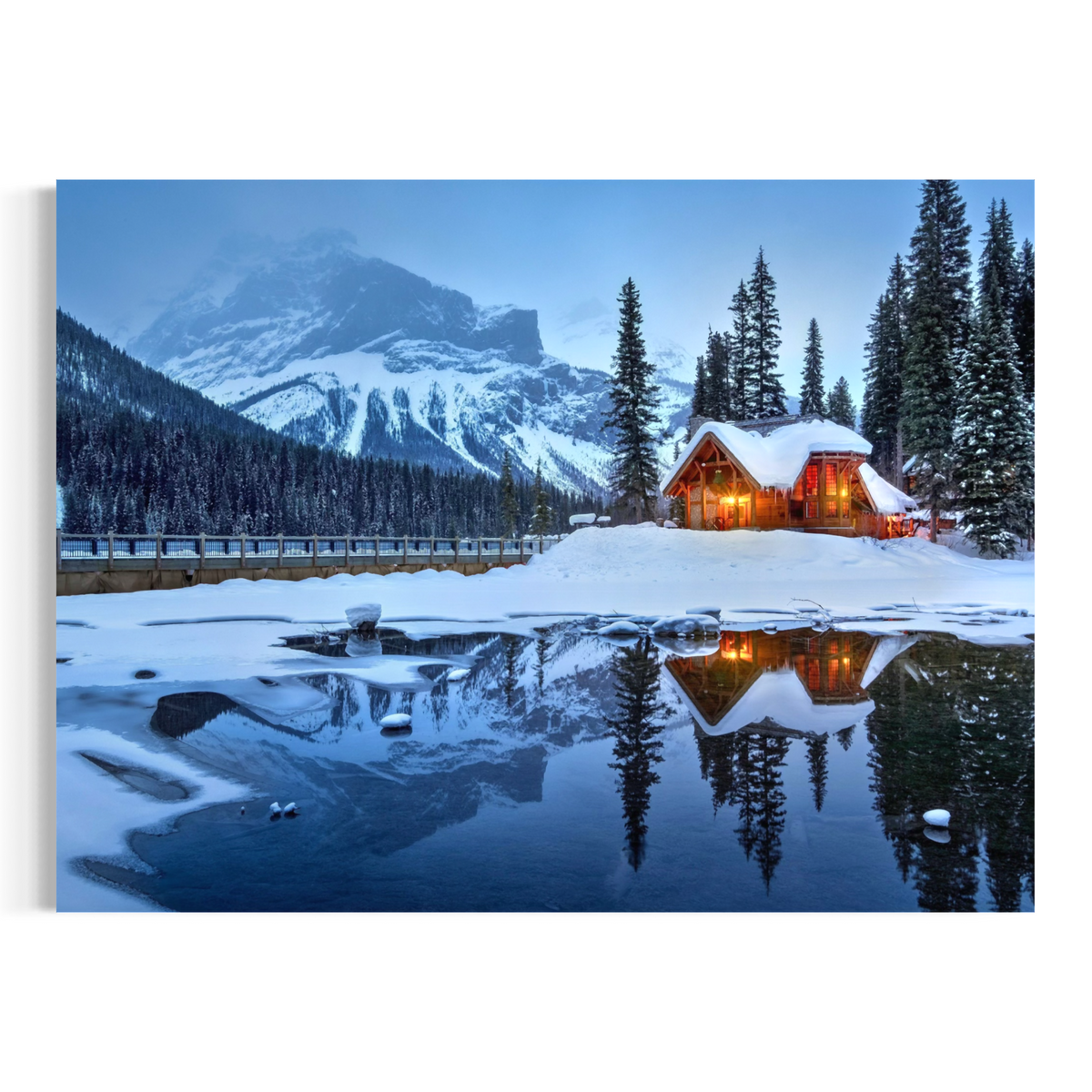 A scenic winter landscape print showing a snow-covered mountain, evergreen trees, a warmly lit log cabin, and a frozen reflective pond in the foreground.