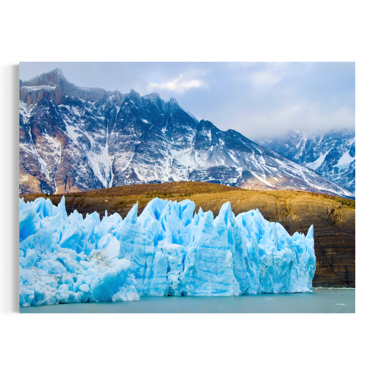 A rectangular wall art print showing a glacier in the foreground with snow-covered mountains and cloudy sky behind it, displayed against a plain white background.