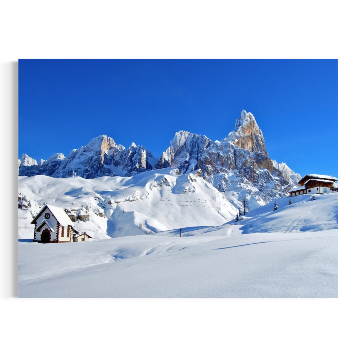 A square wall art print or canvas showing a snowy mountain landscape under a vivid blue sky, with small alpine buildings in the foreground, photographed against a light neutral background.