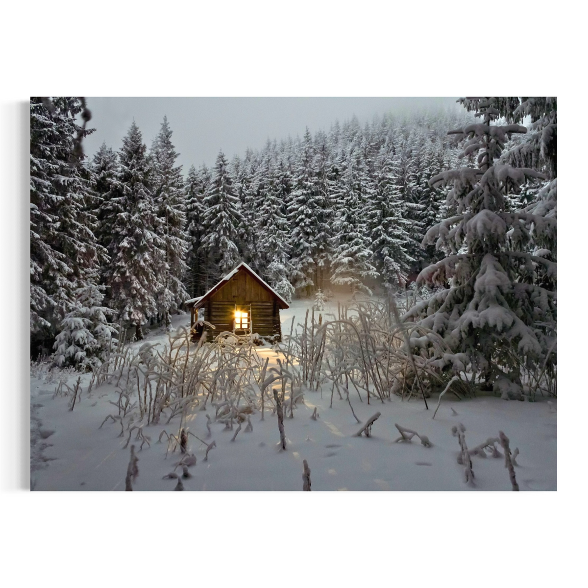 A square wall art print or canvas showing a snowy forest scene with a small cabin lit from within, surrounded by snow-covered evergreen trees and tall dry grasses in the foreground.