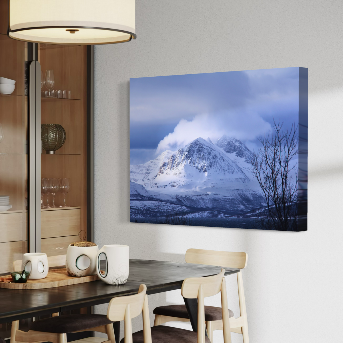 A canvas wall art print depicting a snowy mountain under a blue cloudy sky, displayed on a light gray wall in a modern dining room setting with a wooden table and chairs.