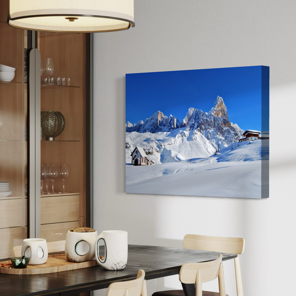 A canvas wall art print depicting a snowy mountain landscape under a vivid blue sky, displayed on a light gray wall above a dining table in a modern interior setting.