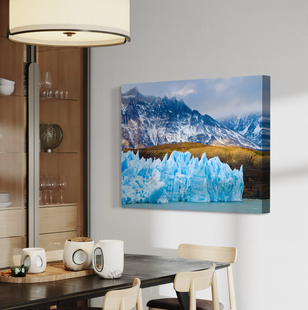 A canvas wall art print showing a dramatic mountain landscape with bright blue ice formations in the foreground, displayed above a dining table in a modern interior setting.