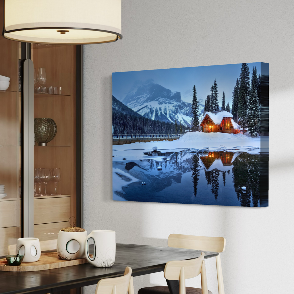 A canvas wall art print showing a snowy mountain cabin reflected in a blue lake, displayed on a light gray wall above a dining table in a modern interior.