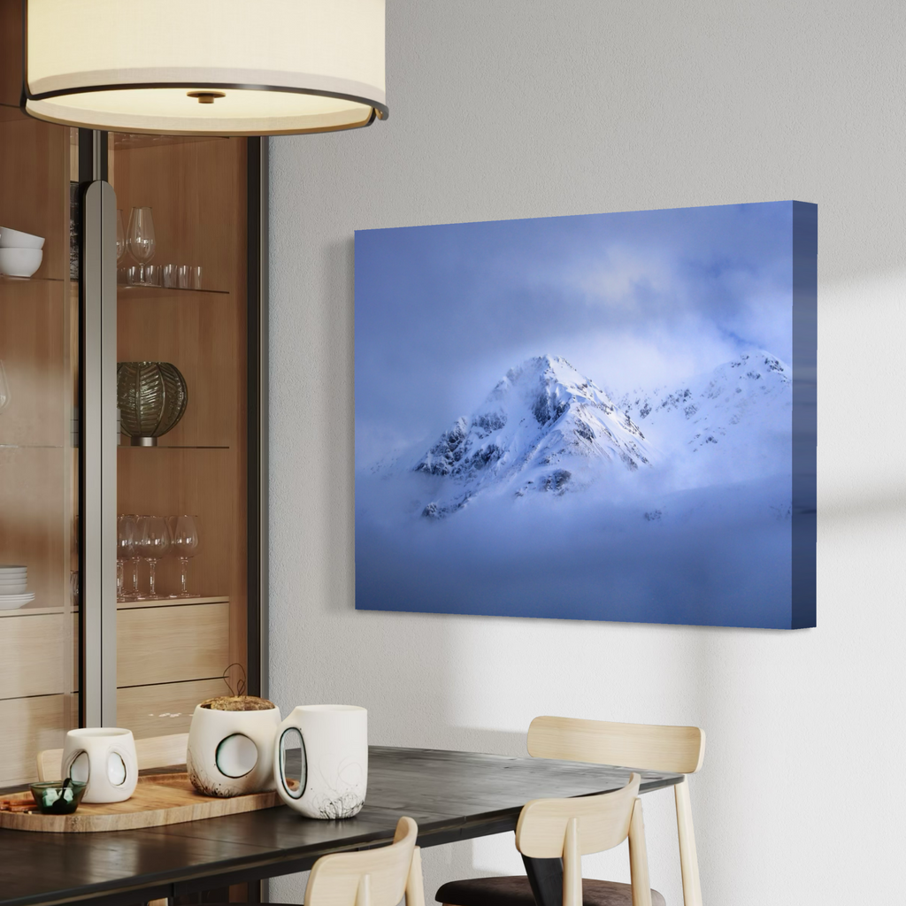 A canvas wall art print depicting a snowy mountain peak rising through blue mist, displayed on a light gray wall in a modern dining room setting.