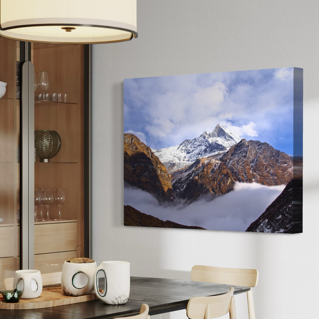 A canvas wall art print showing a snow-capped mountain peak above clouds, displayed in a modern dining room setting above a dark table with white mugs and chairs.