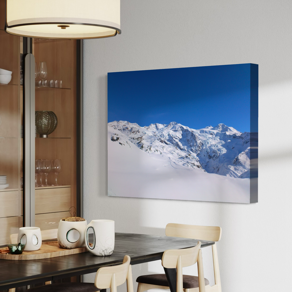 A canvas wall art print showing a snowy mountain landscape under a vivid blue sky, displayed above a dining table in a modern interior setting.