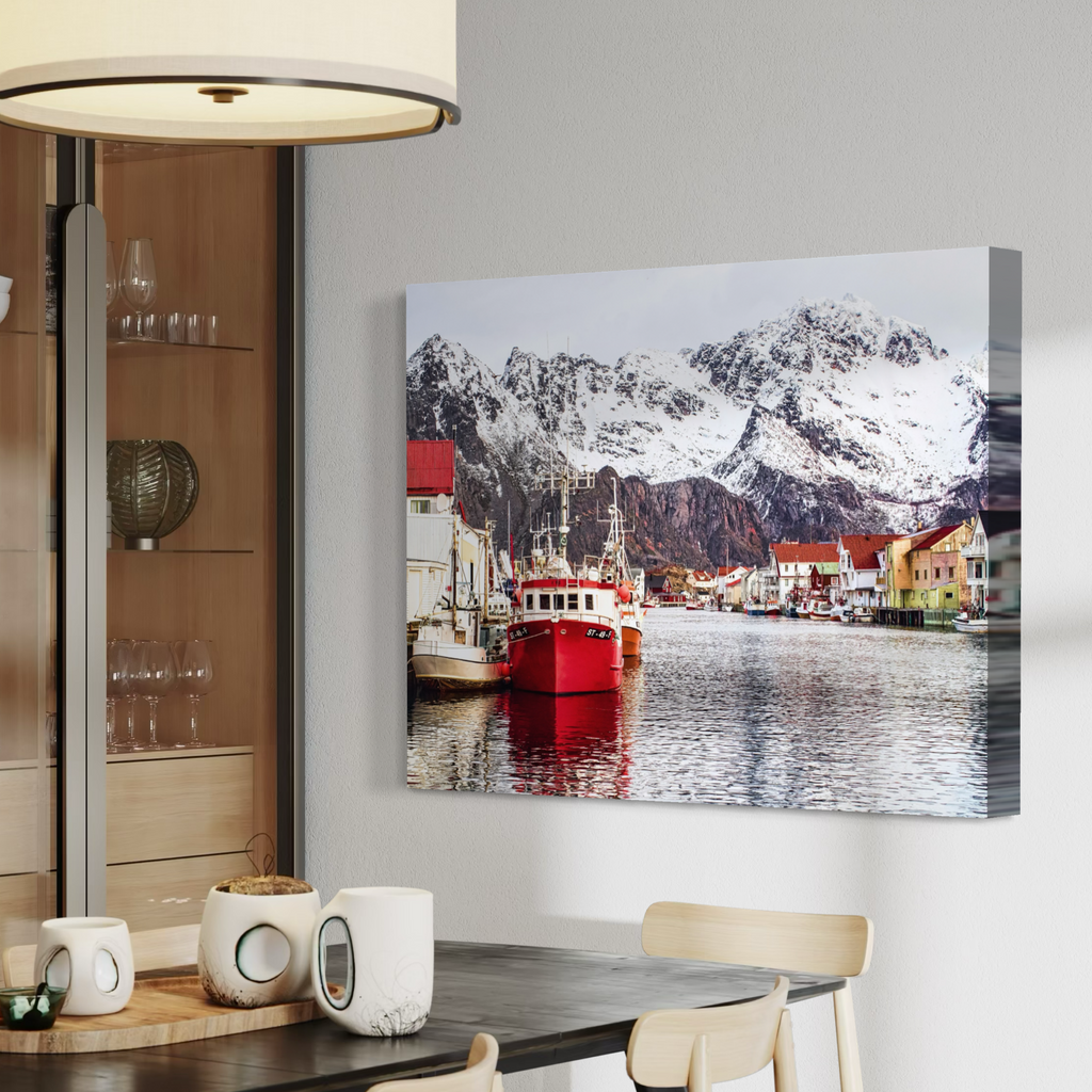 A canvas wall art print showing a snowy mountain harbor with boats and colorful buildings, displayed on a light gray wall above a dining table in a modern interior.